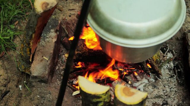 Overhead shot of metal cauldron suspended above active campfire, bright orange flames licking bottom of pot, preparing meal during outdoor adventure, warmth of fire, cozy atmosphere