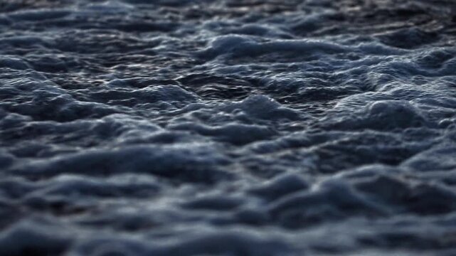 Detailed perspective of dark ocean water with gentle wave motion and reflective surface during twilight. Ripples create abstract patterns, highlighting fluid movement and natural beauty. Slow motion