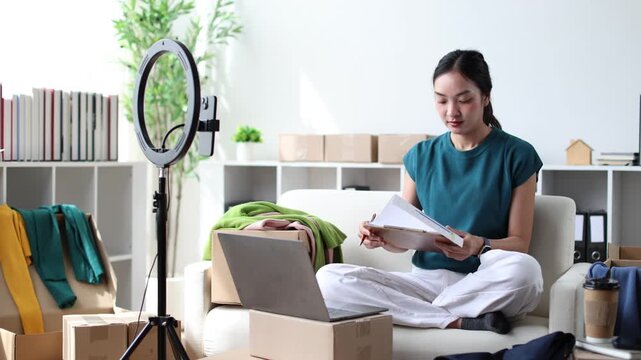 Asian female influencer and sme owner working from home, live streaming and selling clothes on social media using a laptop, a smartphone and a ring light for her online store