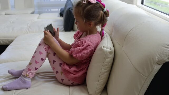 Young girl sitting comfortably on a couch and focusing on a smartphone screen. Bright home interior, childhood, technology use, digital entertainment, communication and modern family lifestyle concept