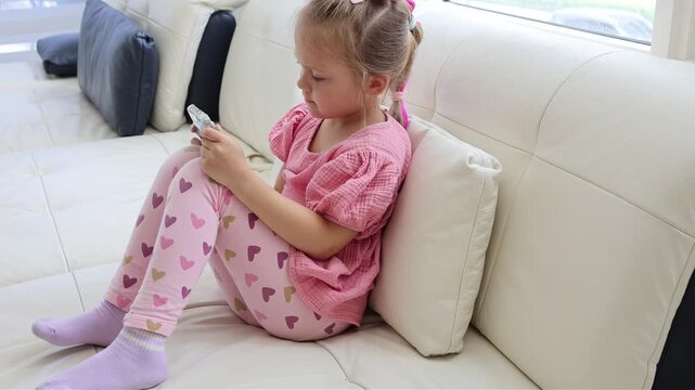 Happy young girl enjoying a smartphone while sitting comfortably on a sofa. Childhood, fun, technology, digital entertainment and modern lifestyle concept.