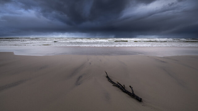 View of a piece of driftwood on a sandy beach under dramatic storm clouds and crashing waves in Debki, Pomeranian Voivodeship, Poland.
