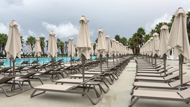 Sun loungers and closed umbrellas by resort pool on cloudy day