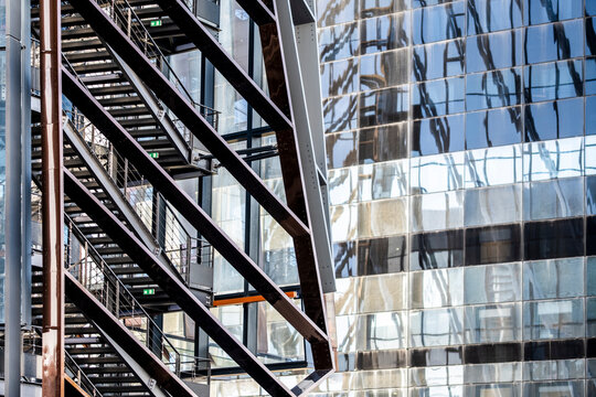 Metal stair structure cuts across urban architecture and a glass facade, where building reflection, window pattern and construction detail create layered exterior lines