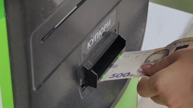Close-up of a hand inserting a 500 Ukrainian Hryvnia banknote into a payment terminal bill acceptor. The process illustrates cash digitalization and automated financial services.