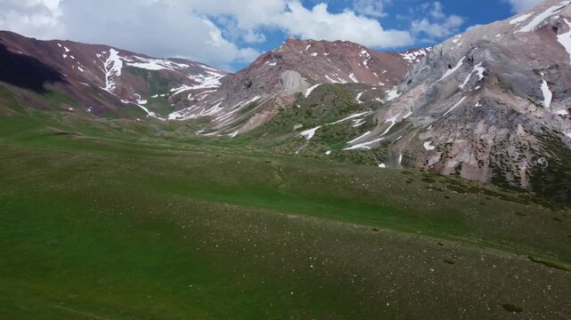 Aerial panning shot showcasing a vast green highland plateau stretching toward rolling red-rock mountain ridges with scattered snowfields in Sairam-Ugam National Park, Kazakhstan. 