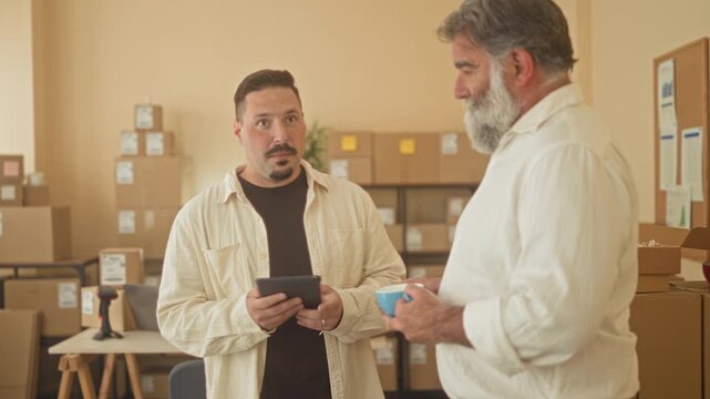 Two men discuss parcels while one holds a tablet and gestures in a packing building; collaboration planning.