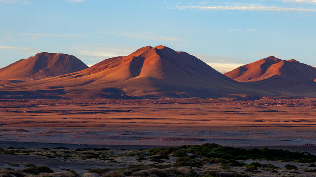 View of reddish volcanic mountains rising above the arid Atacama desert landscape under a soft blue sky at sunset Toconao, Antofagasta, Chile.