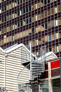 Exterior stairs and metal railing sit against layered urban architecture, where facade grid, building texture, shadow and structure create a compact city detail