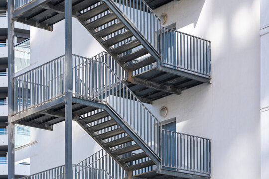 Exterior metal stairs connect white facade levels in urban architecture, with railing, landing, shadow and building structure arranged in repeating lines
