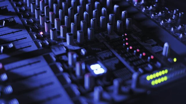 Revealing a professional audio mixer surface with numerous knobs, faders, and blinking lights. The scene is illuminated by cool blue lighting in a dark recording studio environment.