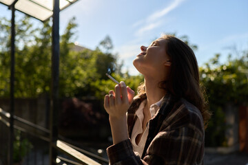 A woman stands outdoors on a sunny day, eyes closed, head tilted back, smoke rising near lips. Casual lifestyle moment capturing reflection, calm, and self care © SHOTPRIME STUDIO