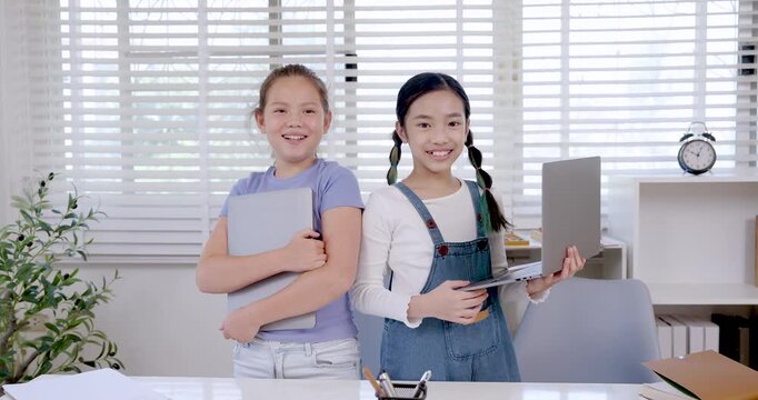 Asian preteen girl holding laptop and caucasian preteen holding notebook smiling together during classroom teamwork collaboration project education research digital learning assignment slowmotion