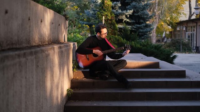 Young European man in glasses tuning guitar before performance in park sunny summer day sunset

