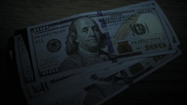 Close-up view of a light beam scanning over a stack of hundred dollar bills on a dark wooden table. The moving spotlight effect dramatically reveals the currency details and wealth concept.