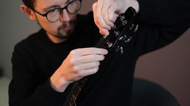 Young handsome talented guitarist in glasses replacing strings on guitar tuning it

