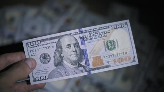 Close-up shot of a hand holding a one hundred dollar bill over a pile of cash in darkness. The flashlight beam reveals banknote details creating a high contrast dramatic atmosphere.