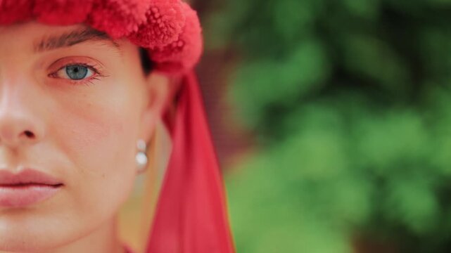 Macro shot of a woman wearing a traditional red floral wreath with ribbons. Focus on the eye and facial details. Blurred green foliage in the background, urban outdoor setting.