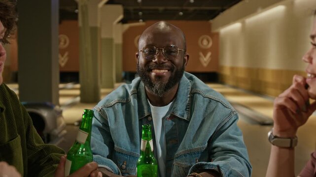 Slowmo portrait of young African American man in denim jacket and glasses smiling at camera while sitting with friends in bowling alley. Beverage advertisements and recreational lifestyle content