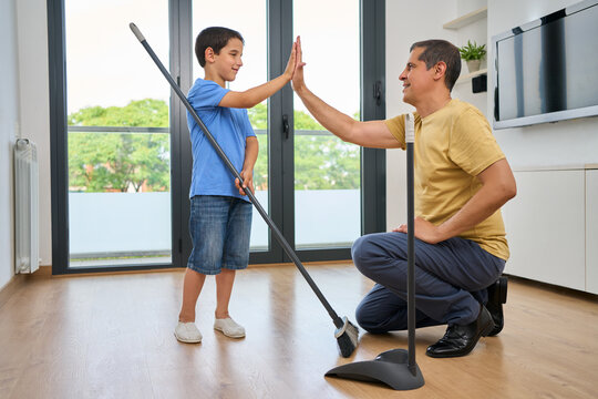 High five while sweeping with father and son in live room,  of home together for weekend chores. Motivation, success or teamwork of man parent and boy child in apartment with broom and mop at home