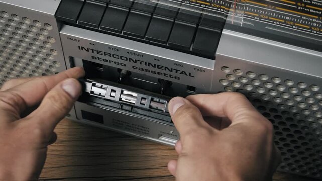 High-angle view of male hands inserting a retro audio cassette with a yellow label into a boombox. The silver door is closed and the start button is pressed to play.