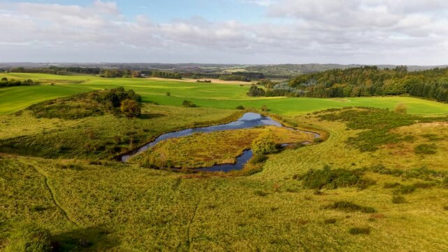 Lush green fields with winding stream and forested hills in Denmark's Jutland peninsula.