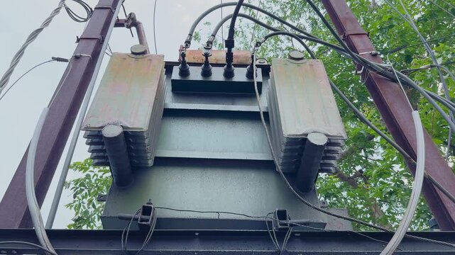 Tracking shot moving left to right across an electrical distribution transformer mounted on a utility structure, showcasing power cables, cooling fins, and urban electricity infrastructure