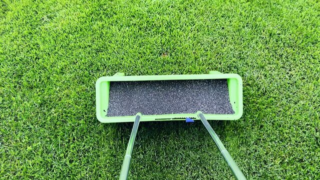 Lawn spreader with black granular fertilizer on green grass under bright sunlight