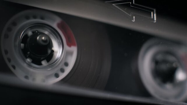 Angled macro view of a transparent audio cassette playing with a spinning white reel. An embossed arrow marking is visible on the shell inside the dark tape deck.