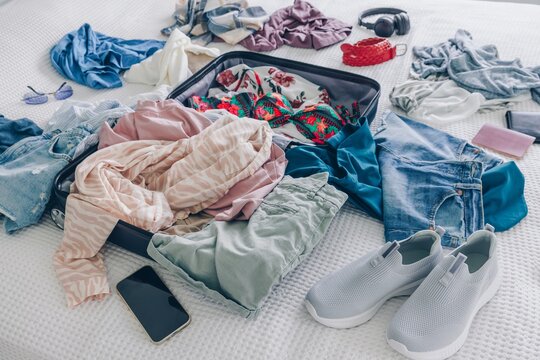 Clothes and Accessories Stacked Around Open Suitcase on Bed with Various Outfits and Items
