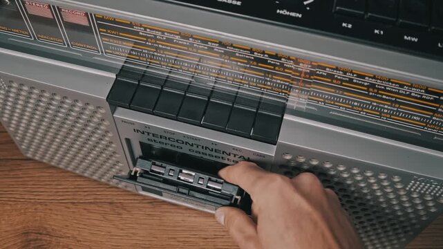 High-angle view of a hand pressing the eject button on a silver retro ghettoblaster. The door opens, and a vintage audio cassette with a yellow label is removed.