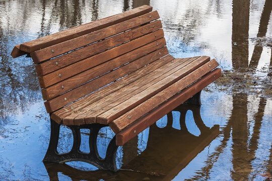 Empty brown stained long wooden sitting bench with black cast iron legs surrounded by flood water in public park flooded by heavy rainfall in spring, Ile des Moulins, Old Terrebonne, Quebec, Canada