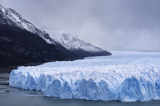 Perito Moreno glacier with shimmering blue ice break edge in Lake Argentino, snow-capped mountains under cloudy sky in the background, Los Glaciares National Park, Santa Cruz Province, Patagonia, Argentina