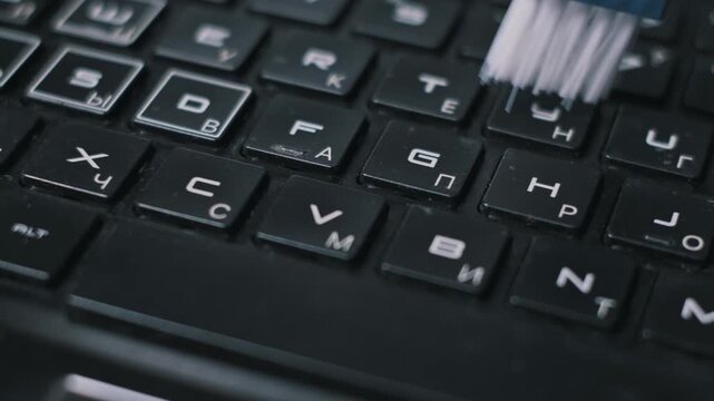 Macro shot of a small blue brush with white bristles cleaning debris from between black laptop keys. The detailed view highlights professional upkeep and sanitization of electronics.