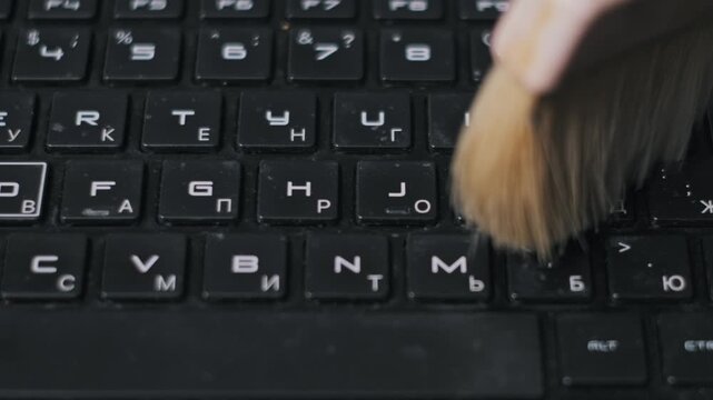Close-up of a wooden brush sweeping dust and debris from a black keyboard with dual alphabet letters. The maintenance shot highlights regular hardware care and office equipment hygiene.