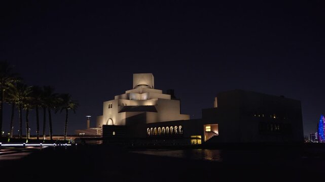 Wide night shot of the Museum of Islamic Art in Doha, Qatar, framed by tall palm trees on the left and distant city skyline on the right, softly illuminated against a deep dark sky.
