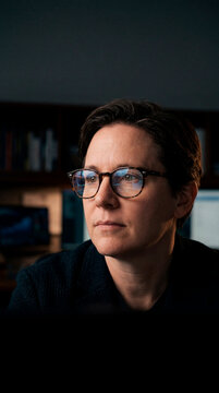 Pensive mature woman wearing glasses reflecting screen data in dark office at night