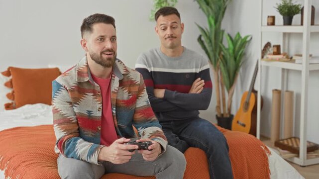 Man holding game controller on bed in bedroom, hands gripping controller while partner watches with arms crossed and smiles; playful intimacy.