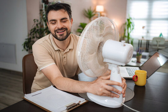 Man working from home enjoying refreshing air from electric fan during summer heatwave