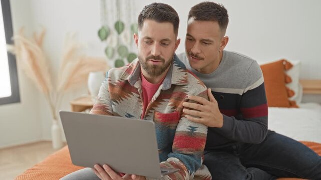 Man couple sitting on bedroom bed with laptop, one man holds laptop while partner places hand on shoulder and leans in; comfort support.