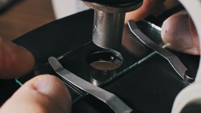 Top-down shot of a scientist using a dropper to apply a clear fluid sample onto a glass slide on a microscope stage. This aids medical experiments or chemical tests.