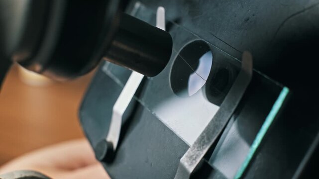 Extreme close-up of a scientist's hand positioning a glass slide with a hair specimen under a microscope objective lens. Metal clips secure the plate for biology.