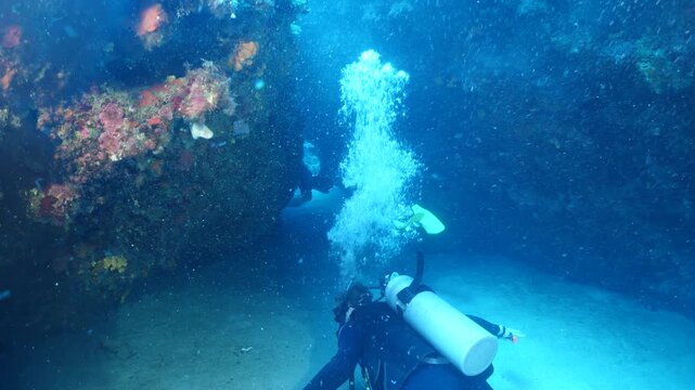  scuba divers exploring around a reef underwater deep blue water big rocks and bubbles ocean scenery 