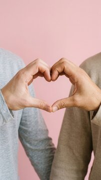 Two people making a heart shape with their hands against a solid pastel pink background