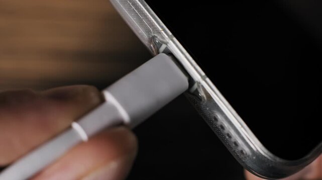 Angled macro perspective of a hand inserting a white Type-C connector into a smartphone side port. The cellular device in a clear bumper case receives current on a wooden surface.