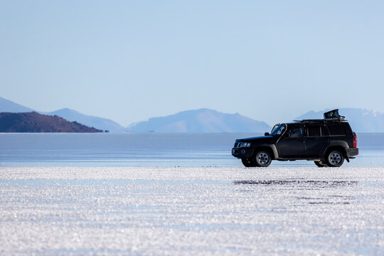 View of a black SUV parked on the white crust of the Uyuni Salt Flat with distant mountains under a clear blue sky in Potosi Department, Bolivia.