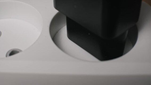 Macro shot of a black Europlug adapter with metal prongs inserting into a white plastic power strip socket. The device connects to the electrical grid to supply household current.
