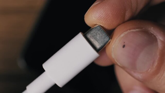 Extreme close-up of fingers holding and inspecting a white USB-C cable connector. The macro detail shows the metal plug reversible design which is a universal standard for devices.