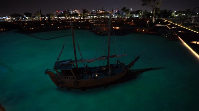 Aerial night shot of traditional wooden dhow boats moored in the illuminated harbor of Katara Cultural Village, Doha, Qatar. Turquoise waters reflect warm lights against a dark city skyline.
