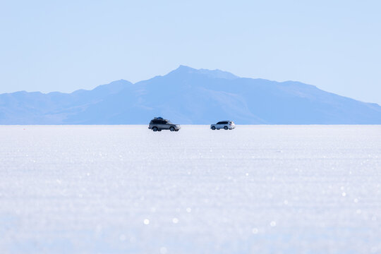 View of two off-road vehicles driving across the vast white expanse of the Uyuni Salt Flat with blue mountains in the background in Potosi Department, Bolivia.